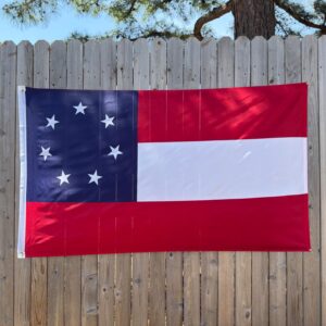 First National Confederate flag, known as the Stars and Bars, hanging on a wooden fence. The flag features a blue canton with a circle of seven white stars, and three horizontal stripes in red, white, and red, displayed in clear daylight with no sunlight showing through the wood slats behind it