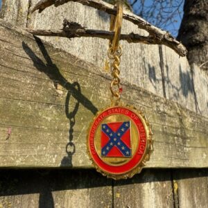 Confederate States of America keychain with gold trim and bold Southern Cross emblem, hanging from a tree branch over a weathered fence.