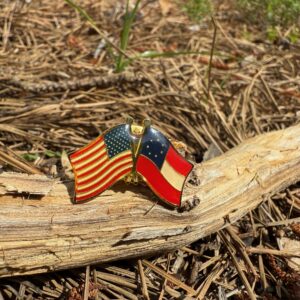 Enamel pin featuring crossed First National Confederate Flag and American Flag with gold trim, displayed outdoors on pine straw and natural wood.