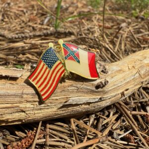 Gold-trimmed enamel pin featuring crossed flags—3rd National Confederate Flag and American Flag—displayed outdoors on pine straw and rustic wood.