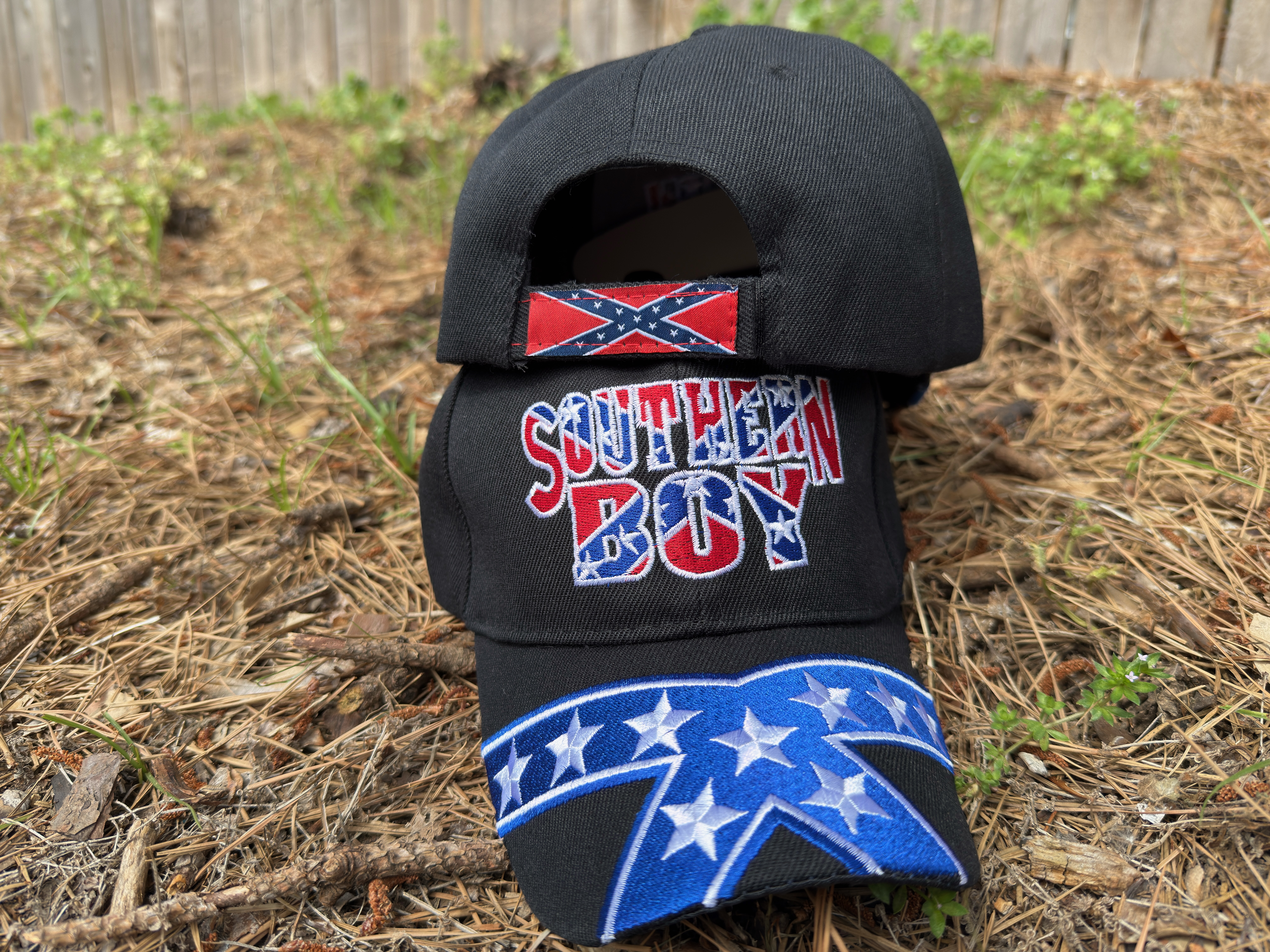 Rear view of Southern Boy Confederate hat featuring Rebel flag on the velcro strap and star design across the bill, resting on a pine-covered ground.