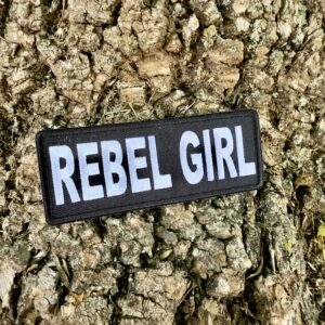Rebel Girl patch with bold white lettering on a black background, displayed on rugged tree bark – 4x1.5 inch iron-on or sew-on Southern biker patch for women.